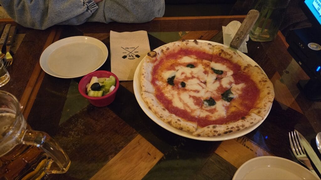A top-down view of a Margherita-style pizza with fresh basil and a mug of draft beer on a wooden table. A side of pickles is also visible
